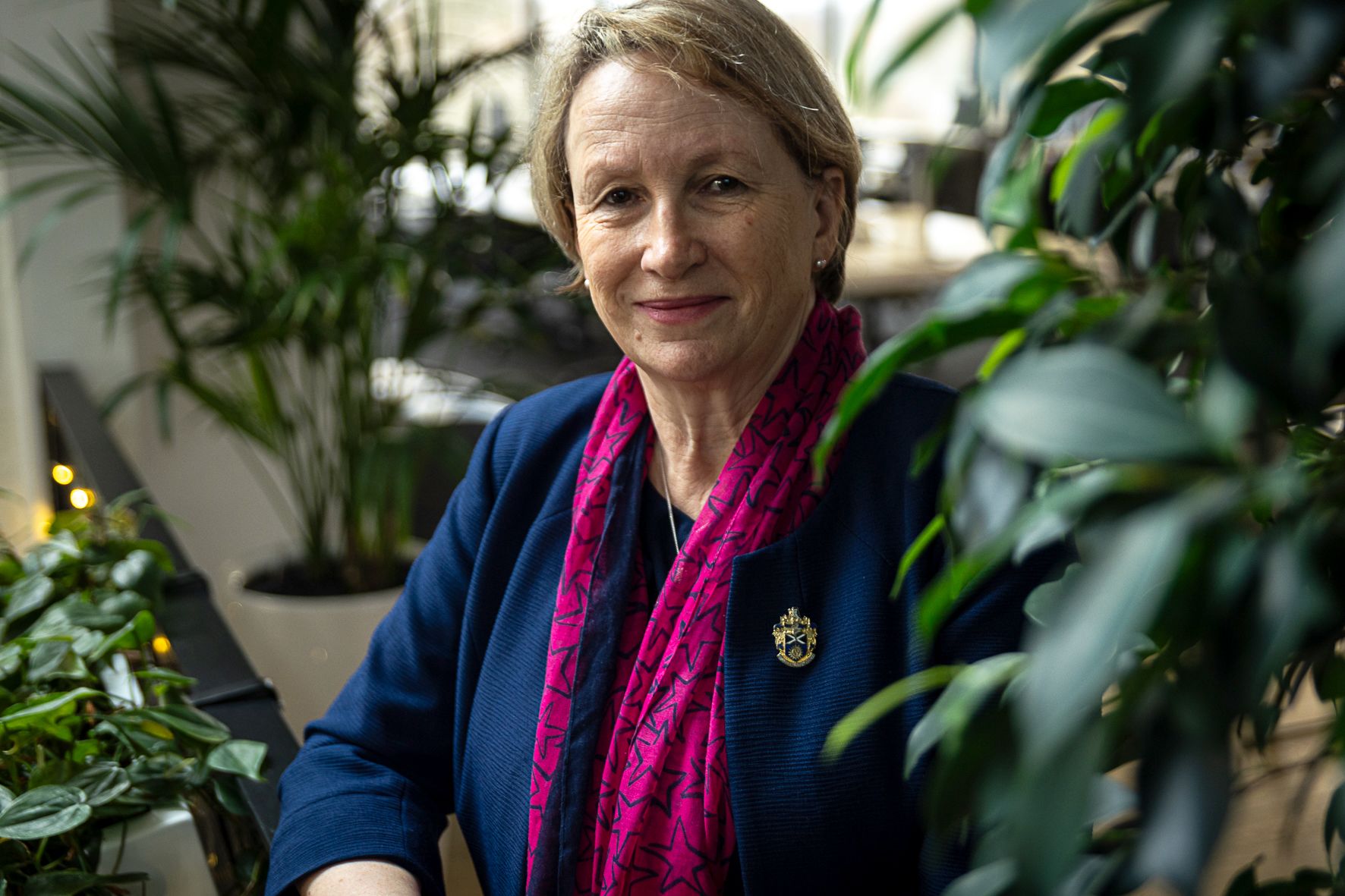 Charlotte Beardmore, pictured smiling in an office with greenery surrounding her. Charlotte is wearing a dark blue blazer, a bright pink scarf, diamond necklace and her SOR brooche