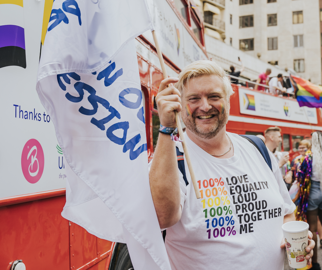 Richard smiling holding a SOR Pride flag, wearing a white t-shirt with pride slogan