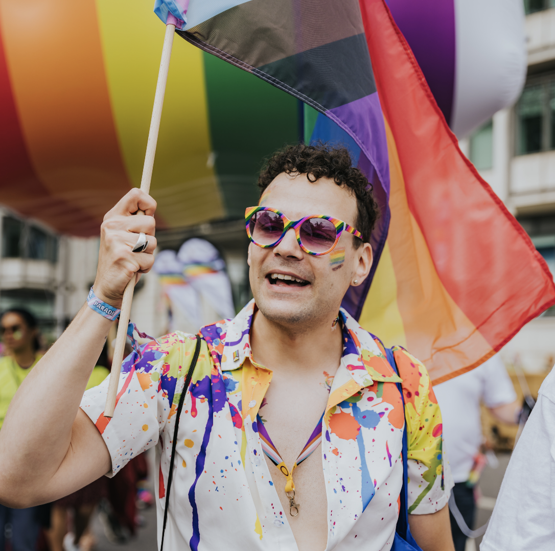 James holding a LGBTQ+ large flag wearing rainbow sunglassed and colourful shirt